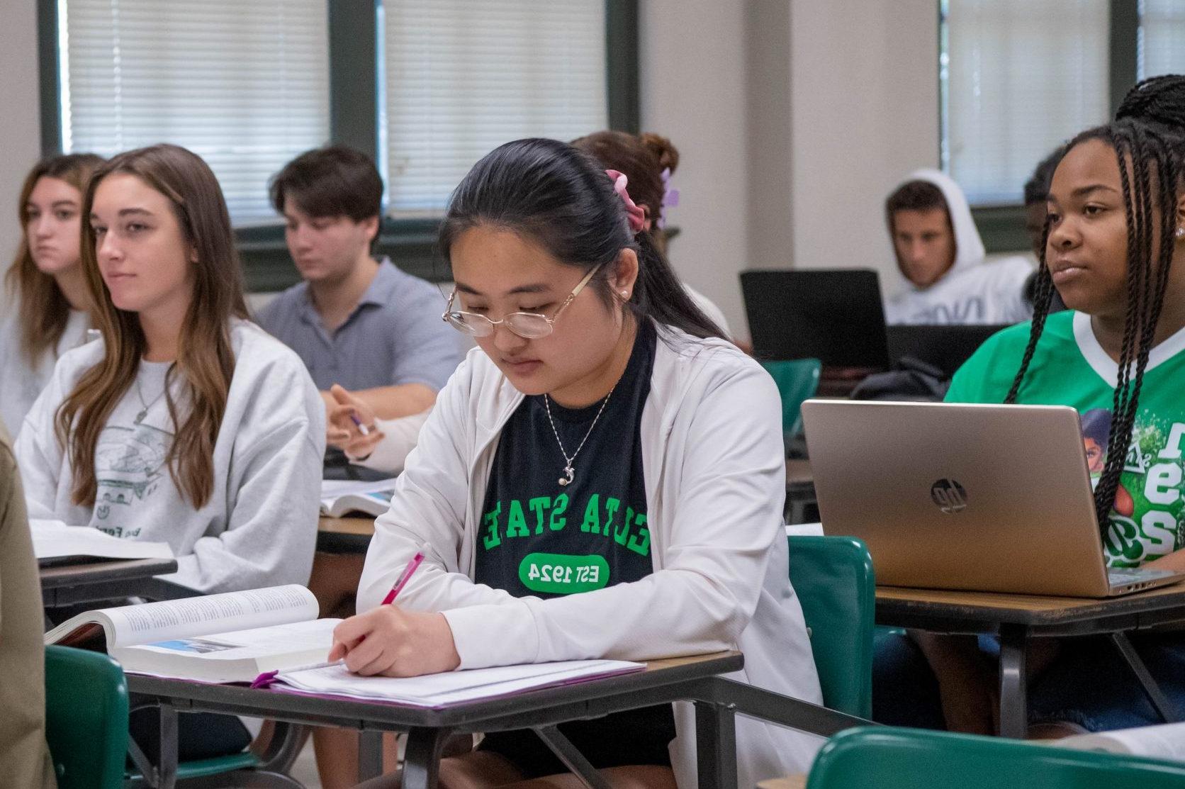 Student sitting at a desk in a full classroom, taking notes on paper with textbook open.