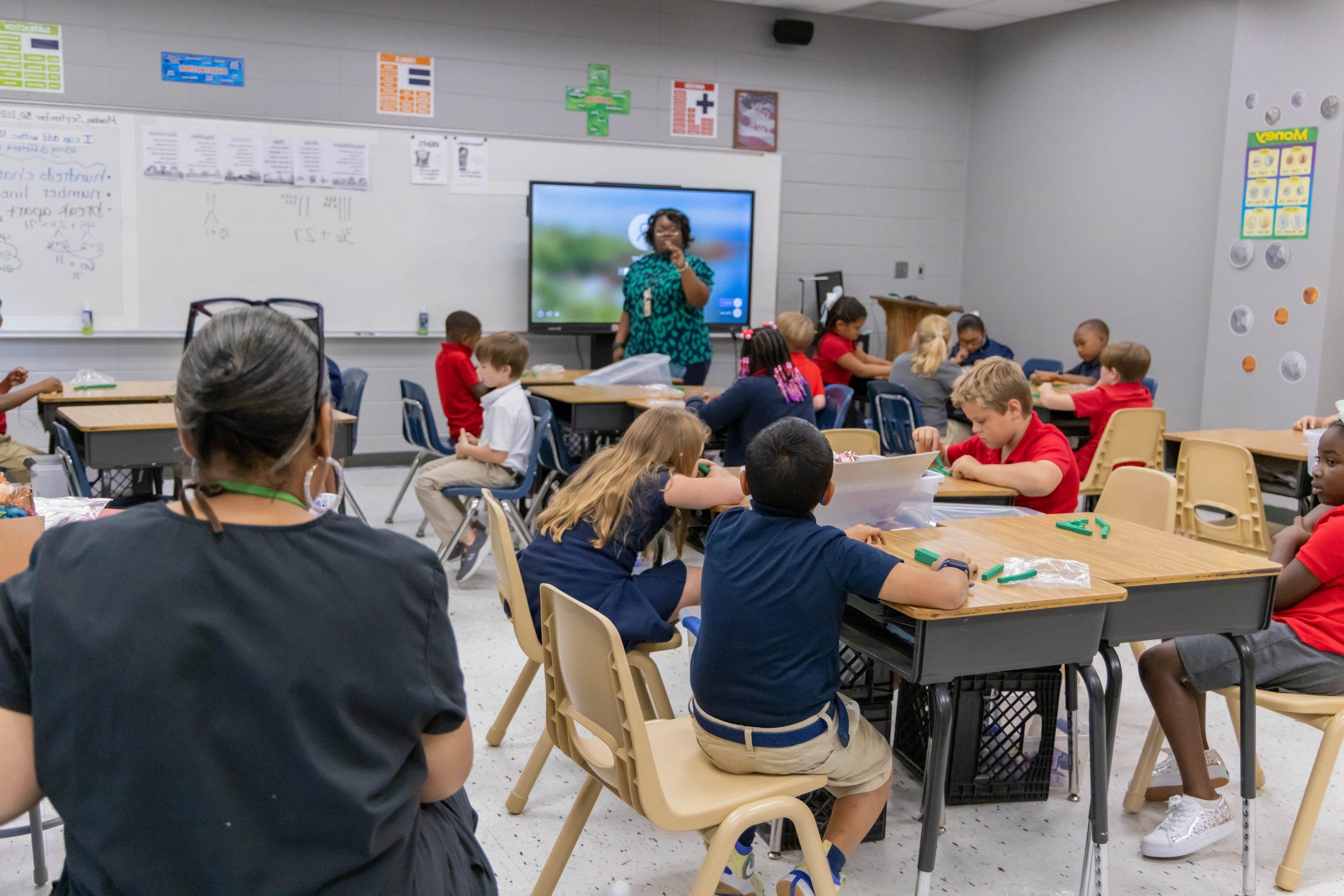 An elementary classroom filled with students sitting at desks, as the teacher teaches class and an administrator observes.